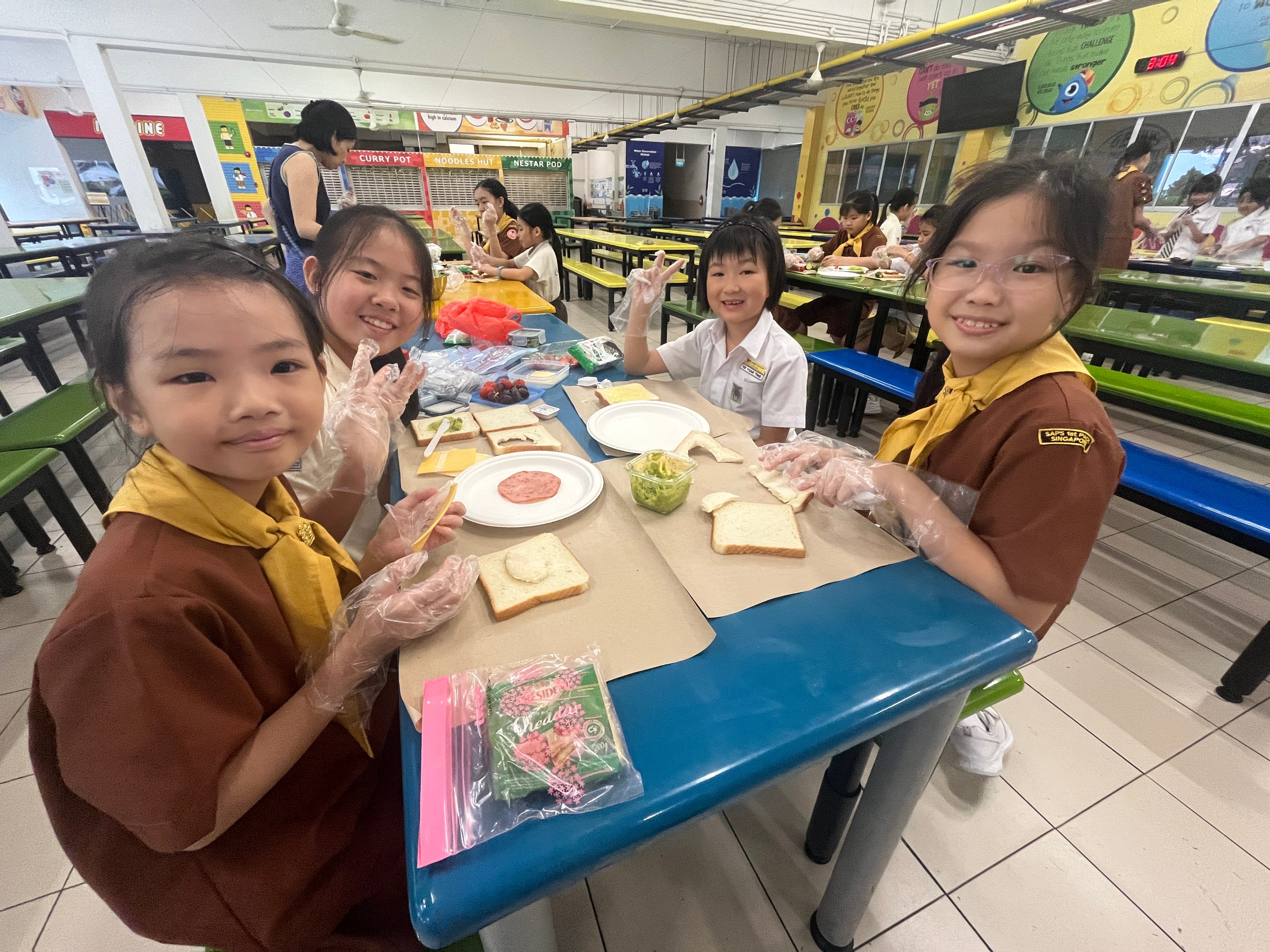 Three smiling schoolgirls in uniforms make sandwiches at a yellow cafeteria table, wearing plastic gloves.